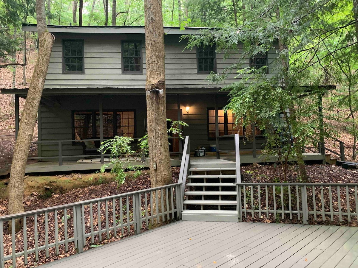The cabin is nestled among trees in a serene forest setting, showcasing a rustic exterior with dark grey walls and green shutters. A spacious deck with a simple railing leads to the entrance, which is illuminated by warm light, enhancing the inviting ambiance.