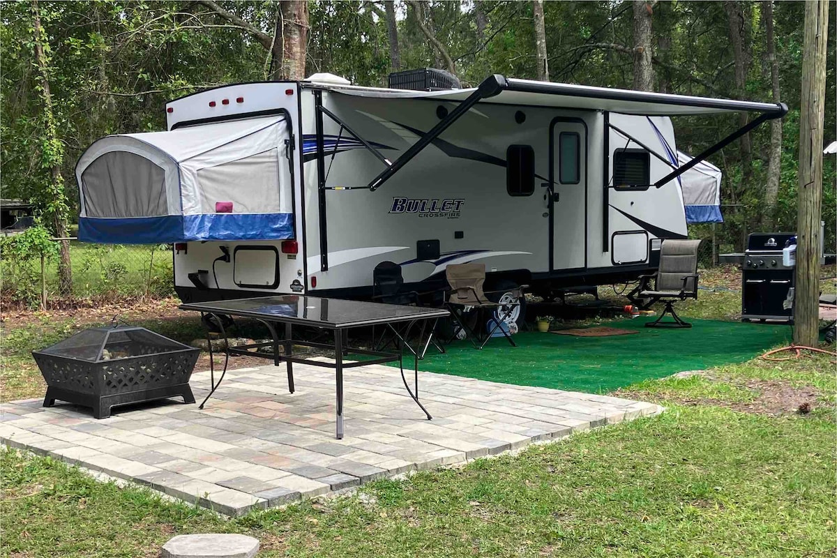A cozy camper is presented with three pop-outs, set on a grassy area surrounded by trees. A patio space features a table and chairs, along with a grill and fire pit nearby. An awning extends from the camper, offering shaded outdoor space.