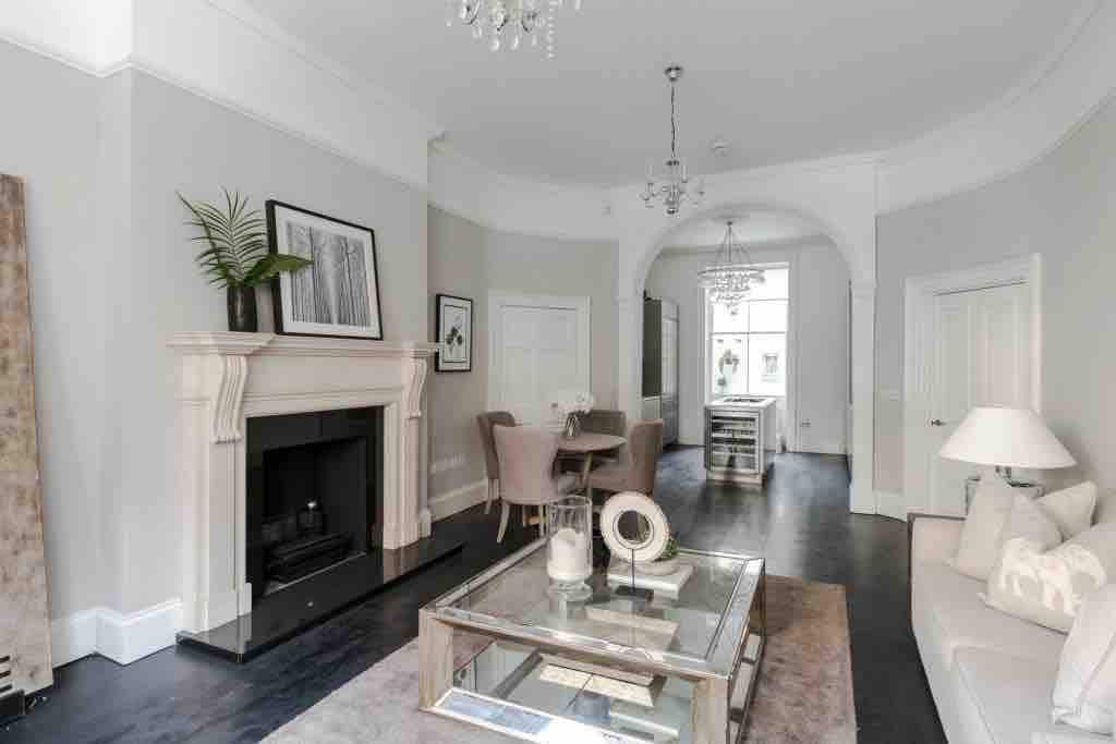 A bright and airy living area is highlighted by a stylish fireplace and elegant furnishings. A modern glass coffee table sits at the center, surrounded by soft seating. Natural light filters through large windows, enhancing the space's inviting ambiance.