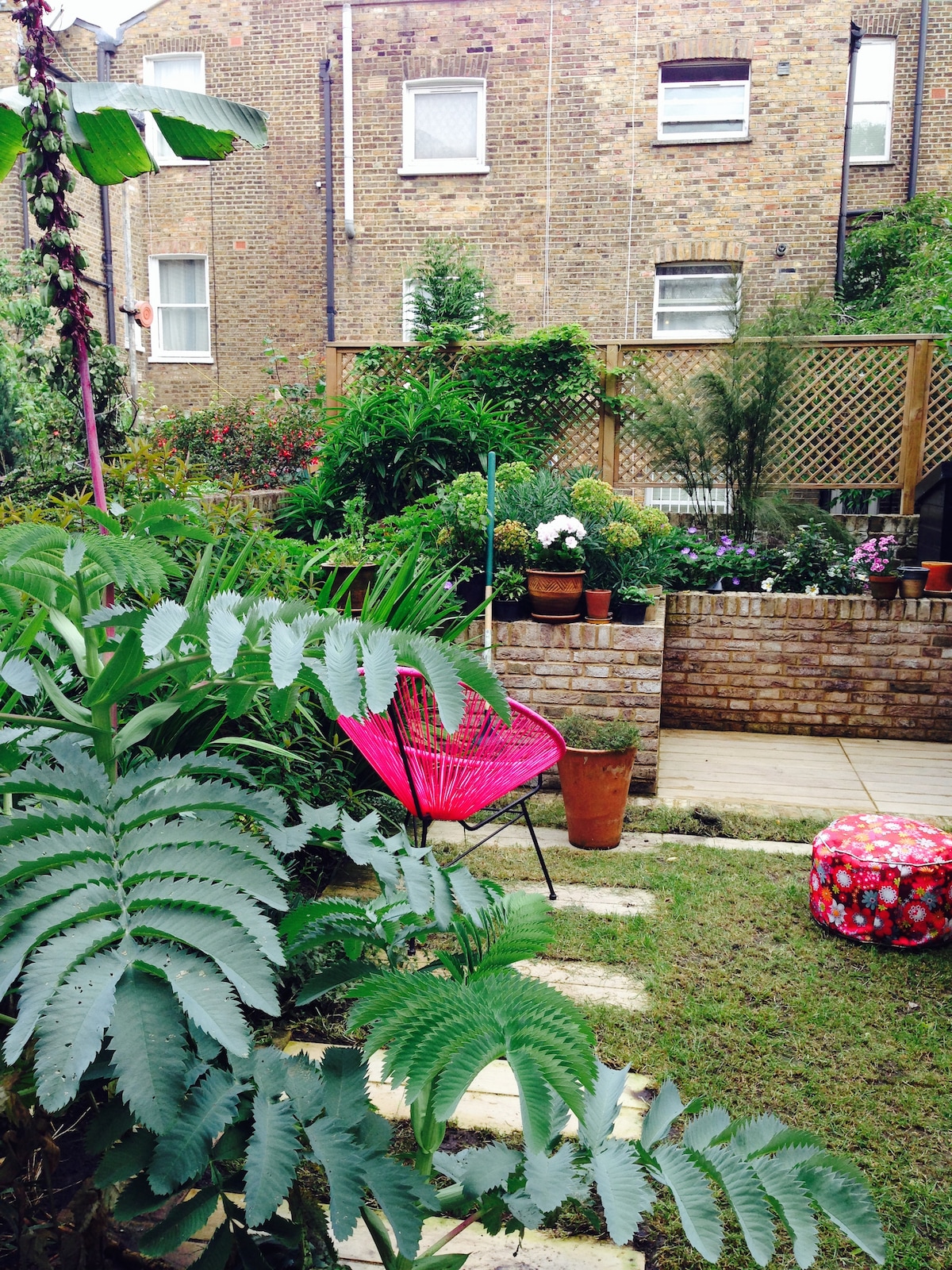 A vibrant garden space is filled with greenery and colorful potted plants. A pink chair is positioned on a stone pathway, surrounded by lush foliage. The peaceful area features a small wall and a variety of plants, creating a relaxing environment.