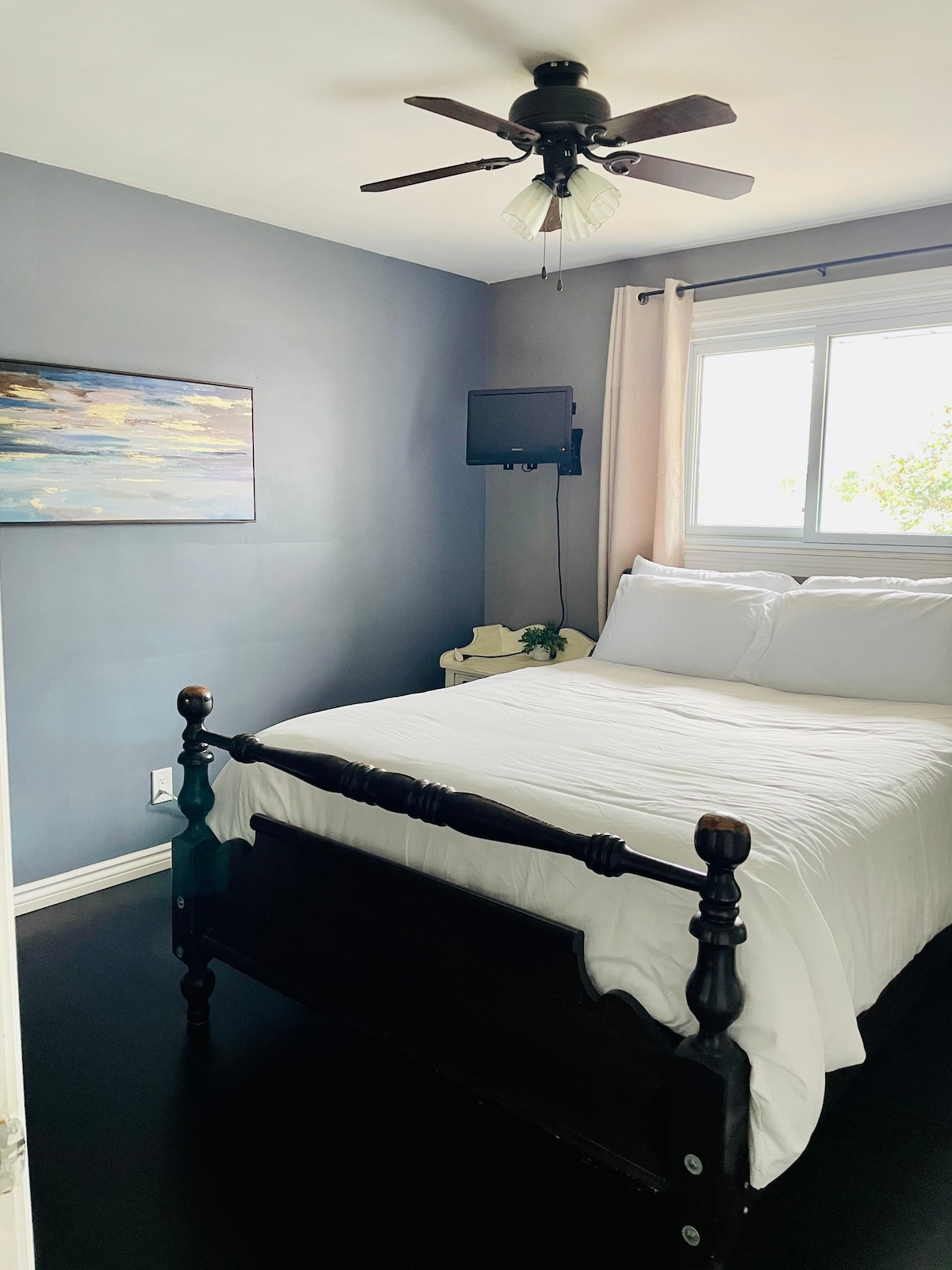 A cozy bedroom features a dark wooden bed with a white comforter and pillows, accentuated by grey walls. A small flat-screen television is mounted on the wall, and natural light fills the room through the window adorned with light curtains.