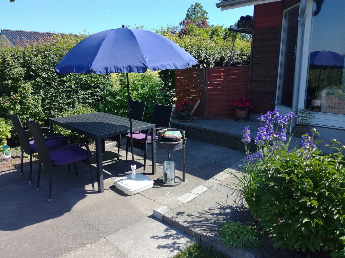 An outdoor seating area features a black dining table surrounded by chairs, shaded by a large blue umbrella. Nearby, colorful flowers add vibrancy to the space, complemented by a small patio area and a well-maintained garden backdrop.