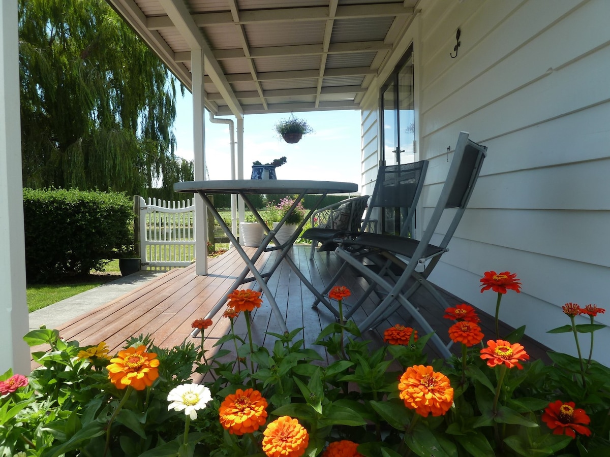 A covered deck area is presented, featuring a round table and several chairs, surrounded by vibrant orange and white flowers. In the background, a lush green hedge and a hanging planter are visible, contributing to a serene outdoor setting.