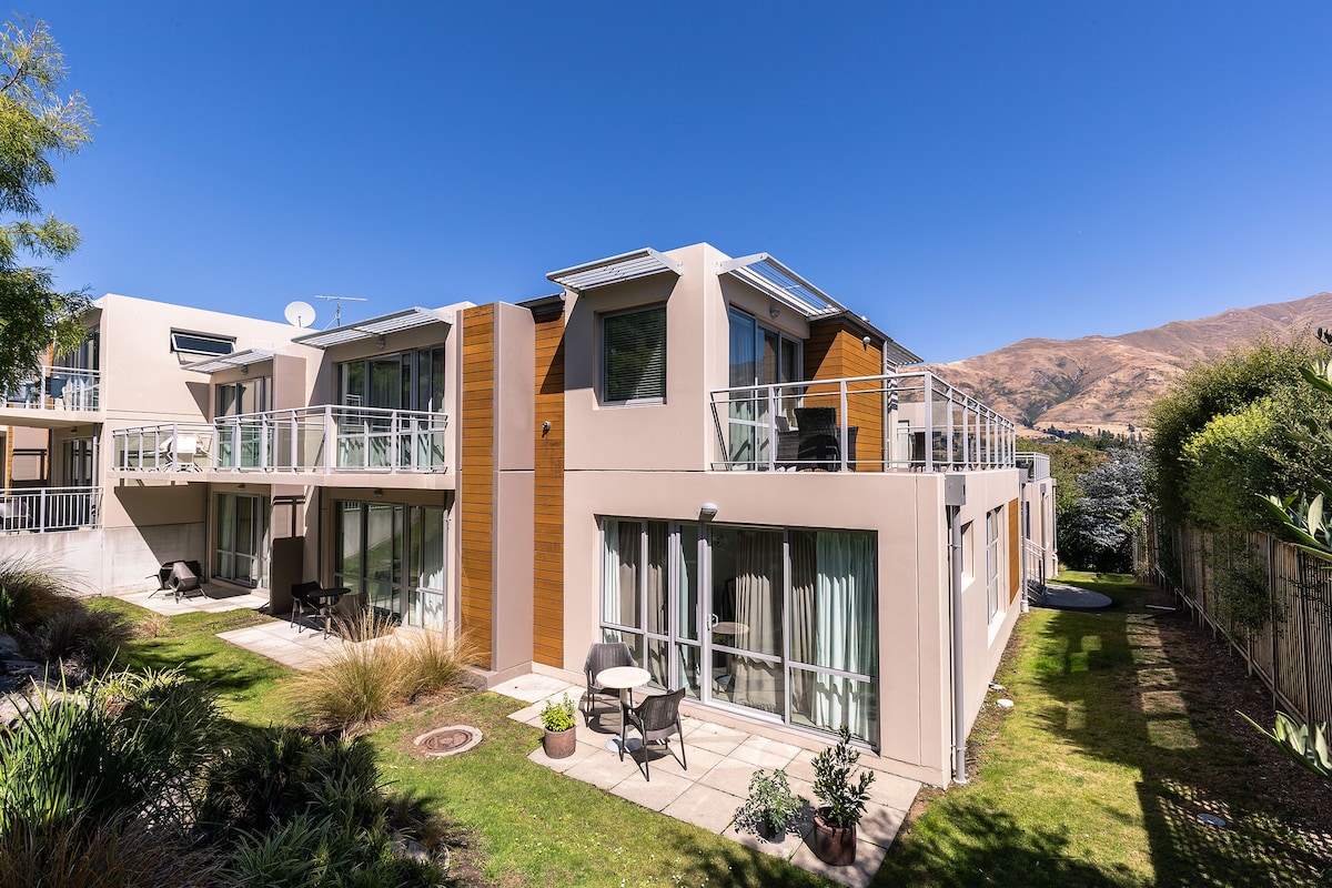 The exterior of the Belvedere Apartments showcases a modern two-story building with a blend of light-colored walls and natural wood accents. A well-maintained garden area is visible, featuring seating on patios and scenic mountain views in the background.