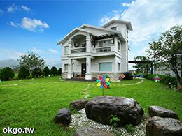 A two-story villa is set amidst a lush green lawn, featuring a welcoming facade with large windows and a covered balcony. Colorful decorations, including a flower sculpture, enhance the outdoor area. Surrounding trees create a serene atmosphere by the grassy space.