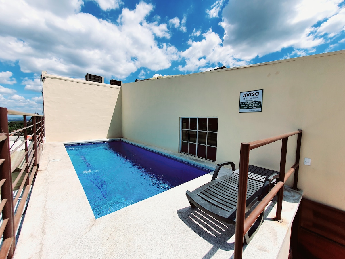 A small outdoor pool is set against a backdrop of clear blue skies and soft clouds. A single lounge chair is positioned nearby, and a safety sign is visible on the wall. The surrounding area is neatly paved, creating a simple and clean space for relaxation.