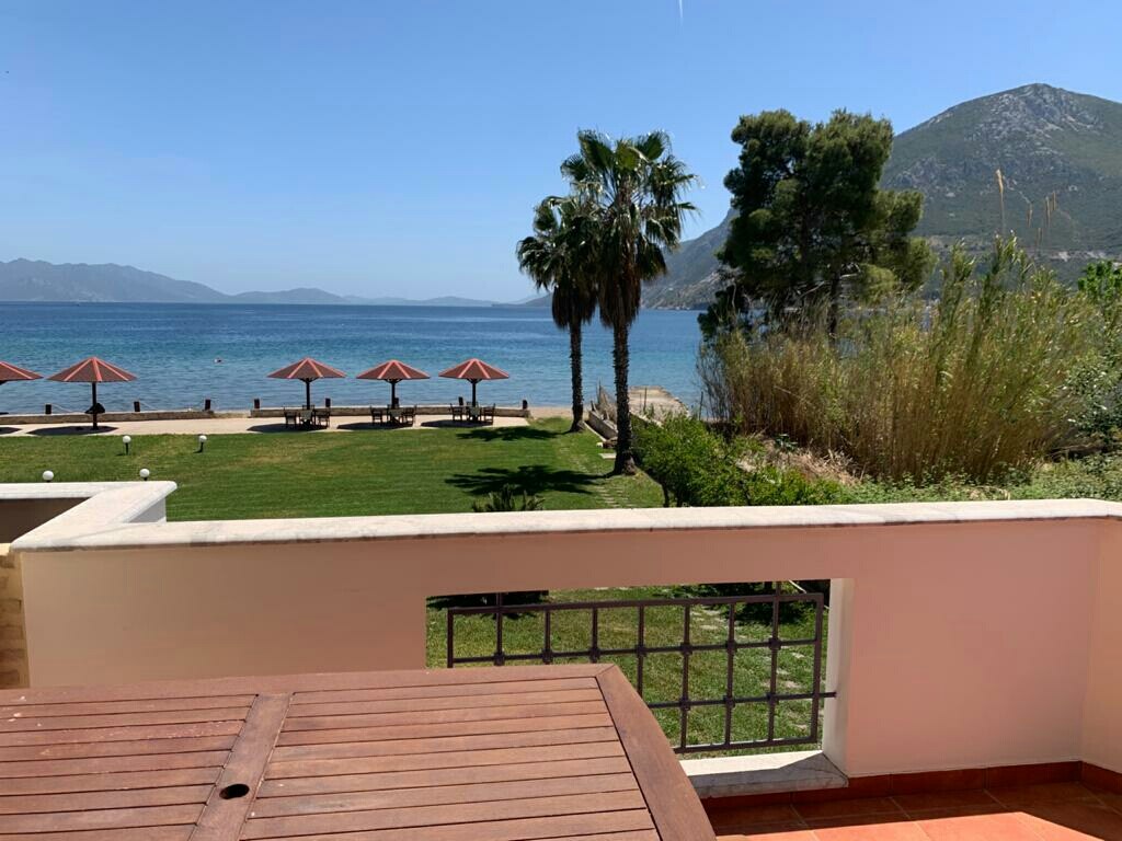 A view of a serene beach from a balcony is displayed. In the foreground, a wooden table is visible. Beyond, several umbrellas are positioned on the sandy shore, with palm trees and greenery surrounding the calm, clear waters. Mountains are seen in the distance under a clear blue sky.