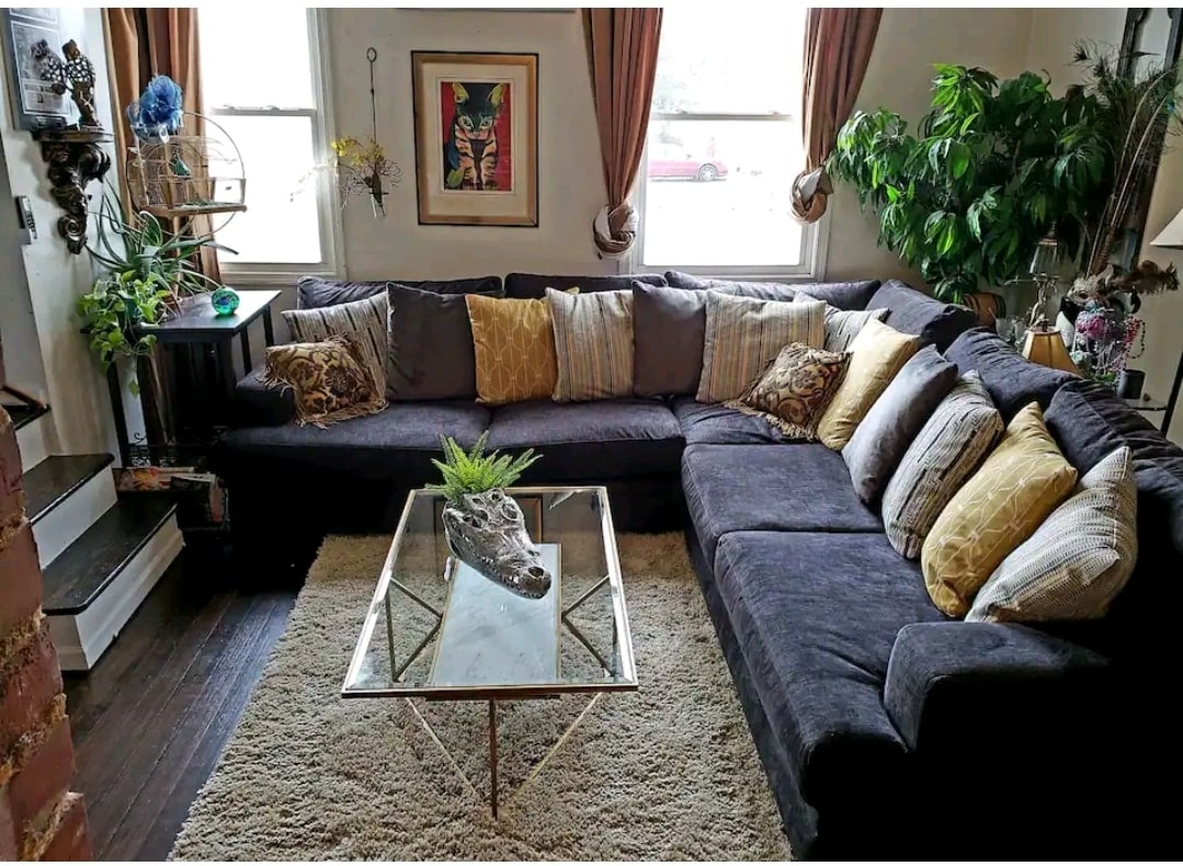 A spacious living area features a large sectional sofa adorned with decorative pillows in varied patterns. A glass coffee table occupies the center of the room, resting on a soft area rug. Natural light filters through the windows, illuminating plants and eclectic wall art.