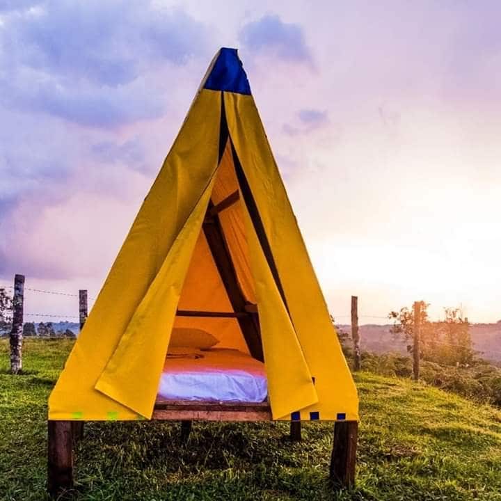 A yellow canvas tent is set on a grassy area, framed by a scenic view of rolling hills and a colorful sky at sunset. The tent entry is open, revealing a spacious bed inside. Wooden posts and a wire fence are visible in the background.