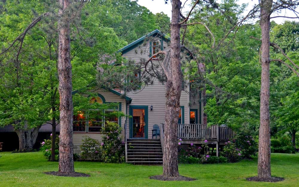A charming two-story house is surrounded by lush greenery, with tall trees framing the view. The front porch features a small seating area, and colorful flowers are visible in the garden. Steps lead up to the entrance, enhancing the inviting appearance of the property.