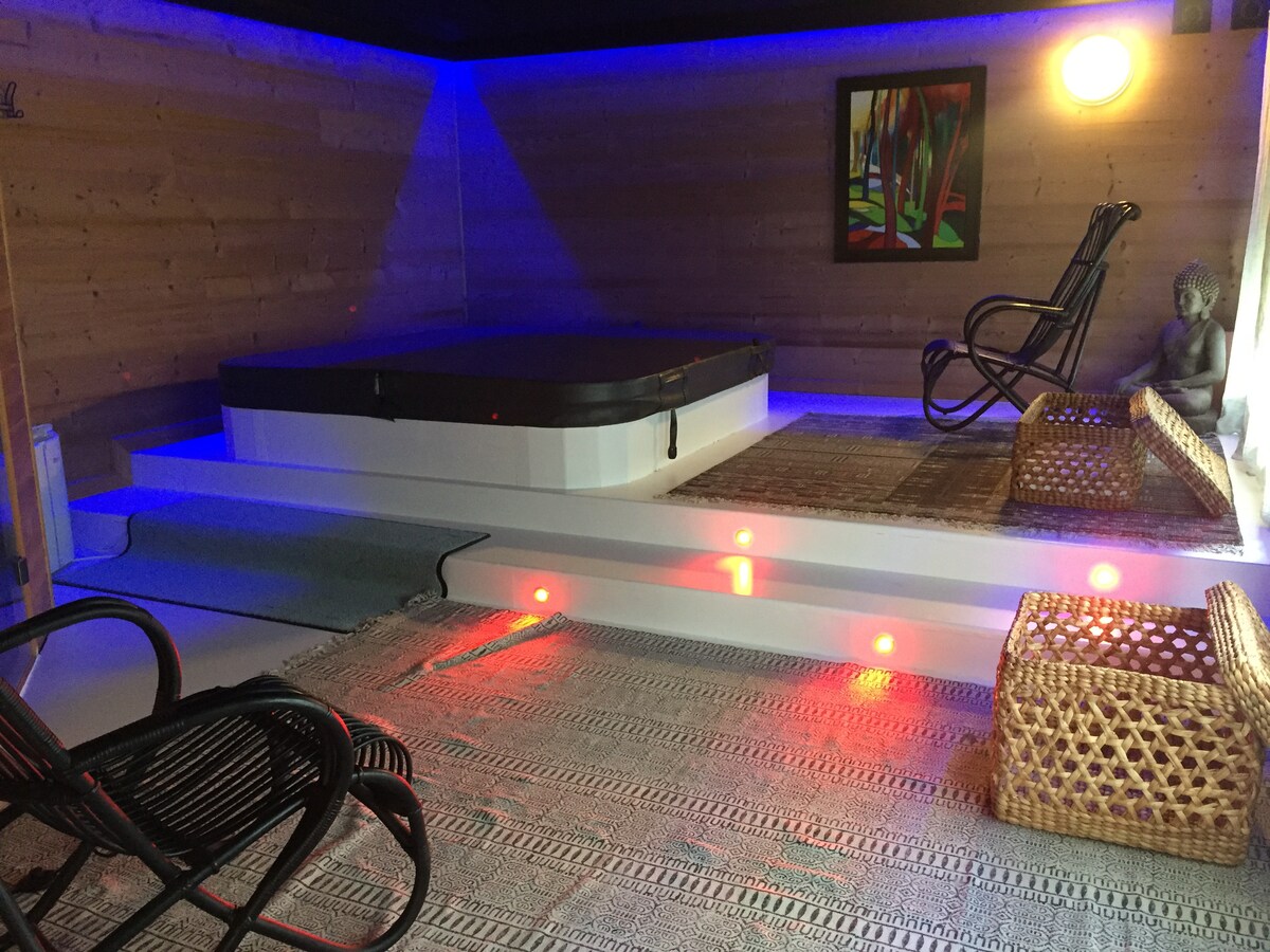 A relaxing wellness area features a hot tub with a dark cover, surrounded by soft lighting. Two wicker chairs are placed on a textured rug, while a colorful artwork is displayed on the wall. A calming atmosphere is created with natural wood paneling.