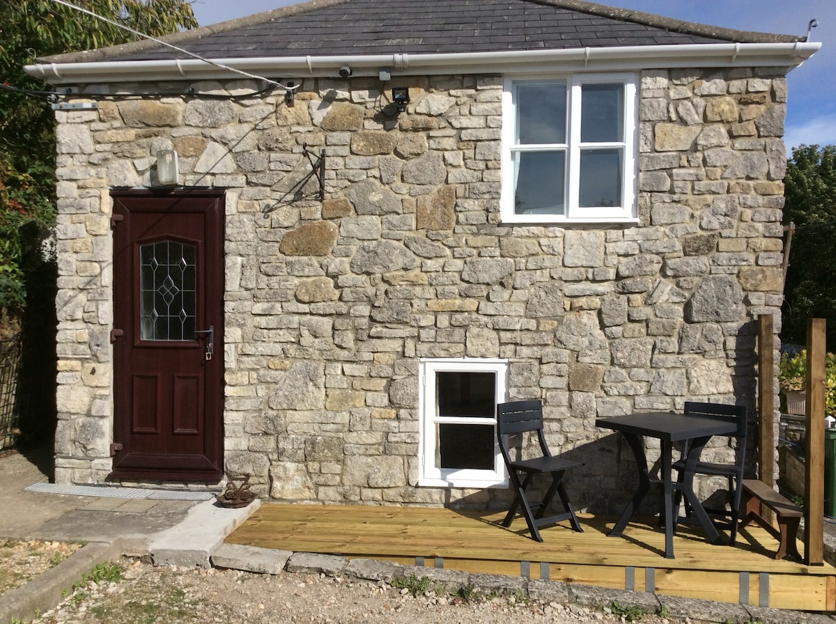 The exterior of a rustic stone guest suite is shown, featuring a dark wooden door with glass paneling and a small window. A wooden decking area with a table and two chairs is positioned outside, offering a welcoming space for relaxation.