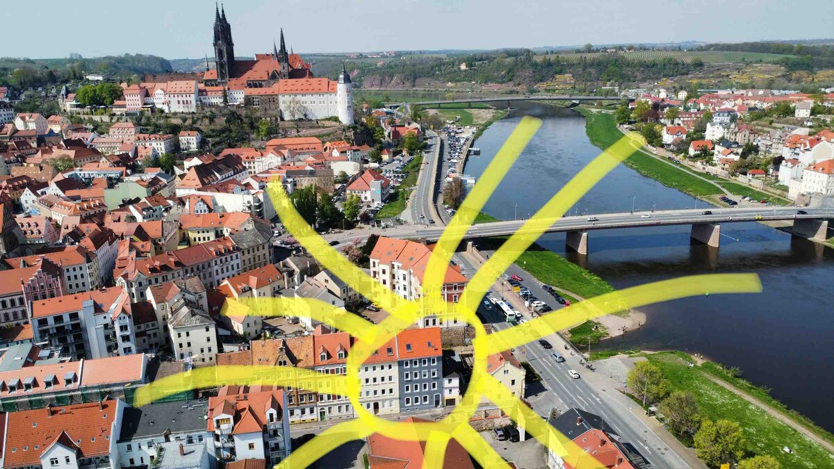 An aerial view captures the vibrant landscape of Meissen, featuring the historic cathedral and scenic river. Red-roofed buildings line the streets below, with a bridge spanning the river, facilitating easy access to the city center.