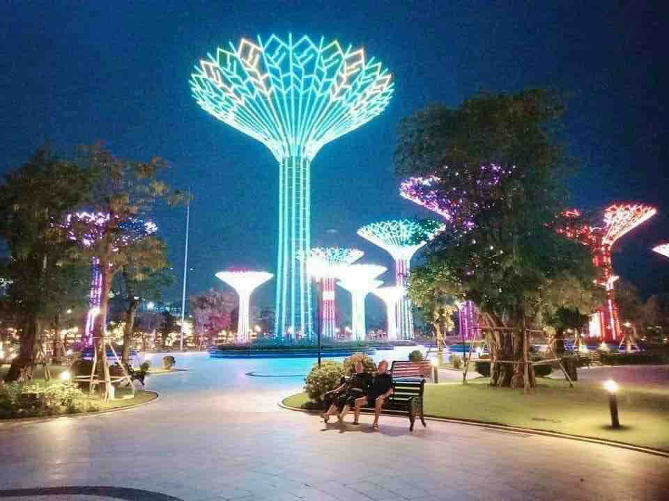 A vibrant park scene is illuminated by towering structures resembling trees, each adorned with colorful lights in shades of blue and green. The well-manicured landscape features paths and greenery, with two individuals seated on a bench, enjoying the nighttime atmosphere.
