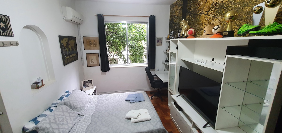 A cozy bedroom is depicted featuring a spacious bed with light bedding and a soft throw. A large window allows natural light, framed by dark curtains. A modern TV unit stands opposite, with a sleek desk and chair nearby. Wall decor includes art and photographs.