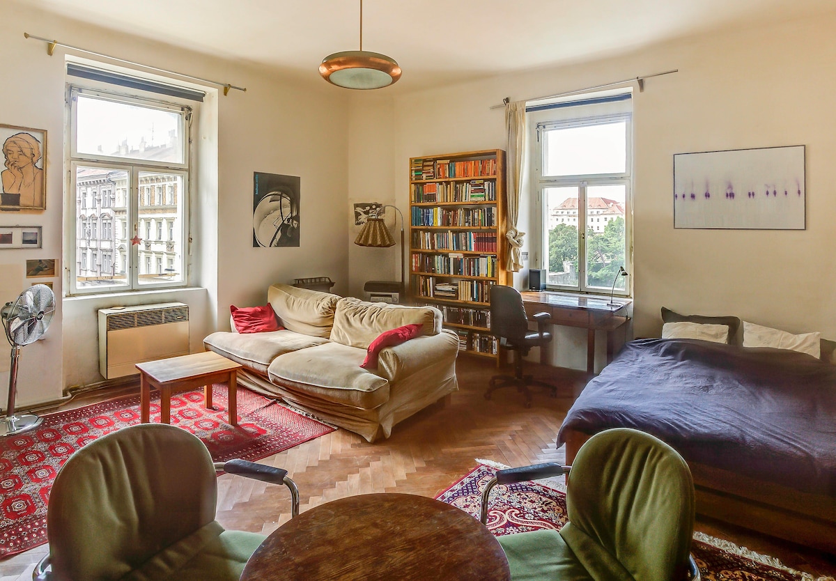 A spacious room is filled with natural light from tall windows, showcasing a blend of seating options including a cozy couch and a comfortable bed. A large bookcase lined with books stands against one wall, while a small desk and a round table with chairs complete the inviting layout.