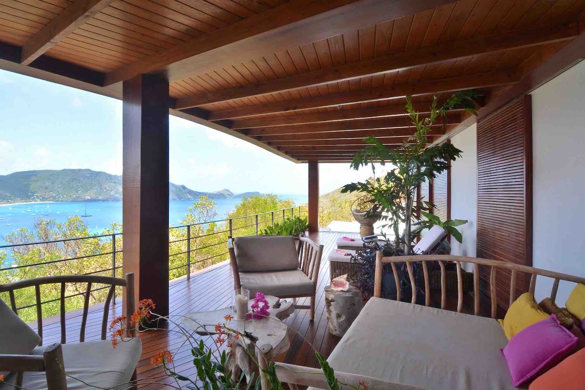 A spacious terrace features wooden beams and a wooden floor, providing unobstructed views of the bay. Comfortable seating arrangements with cushions invite relaxation. Potted plants and decorative touches add a natural ambiance, while the landscape of hills and water serves as a beautiful backdrop.