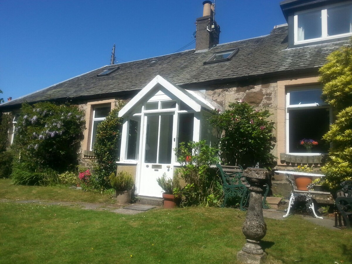 The cottage features a charming entrance porch surrounded by lush greenery, with the facade showcasing traditional stonework. Colorful flowers and plants enhance the garden area, while a mix of outdoor seating is positioned on a neatly kept lawn under clear blue skies.