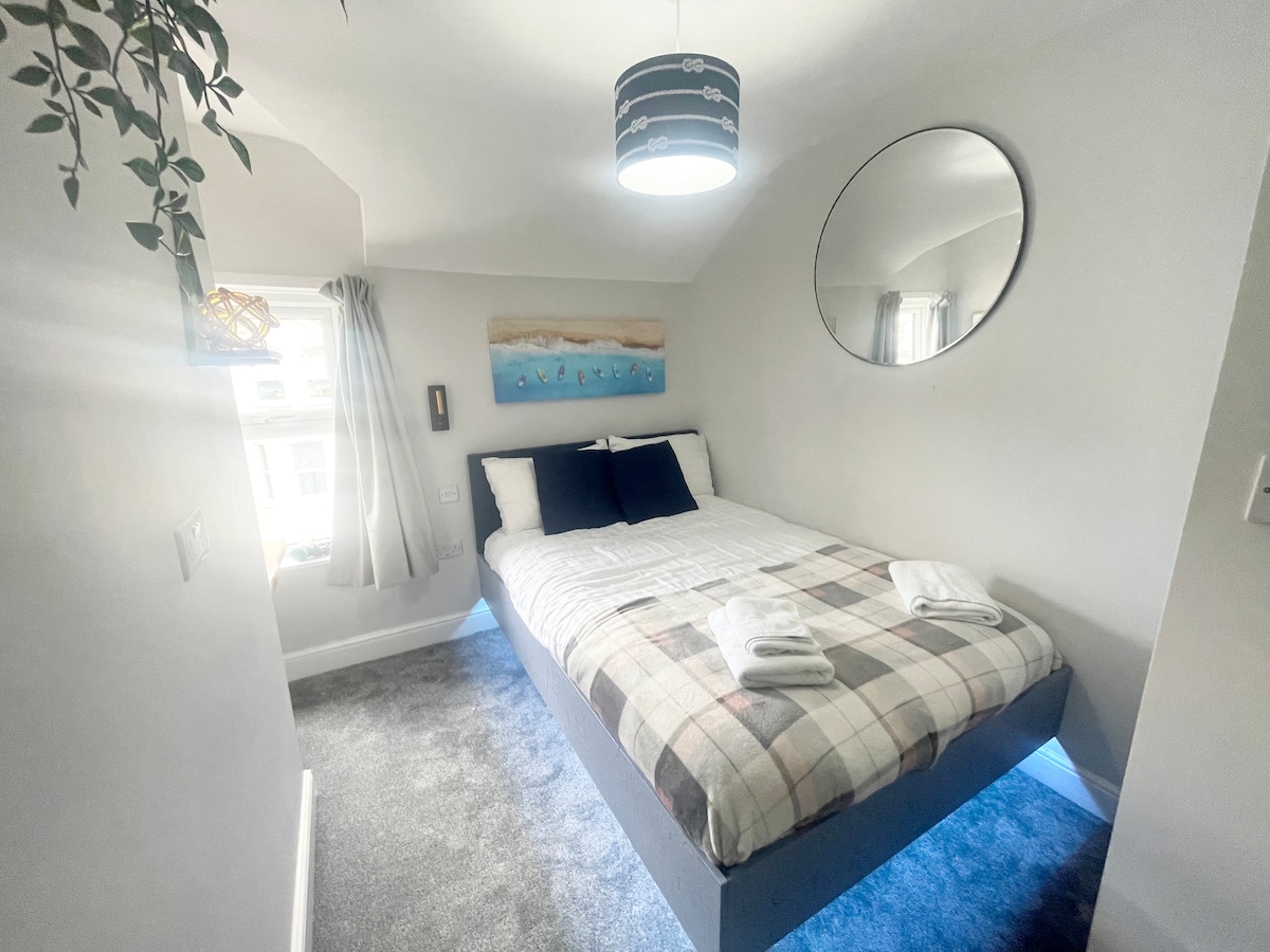A small double room features a spacious bed with a checkered blanket and two neatly folded towels. Soft grey carpet complements the light walls, while a circular mirror reflects natural light from the window adorned with light curtains. A piece of art depicting coastal scenes adds a tranquil touch.