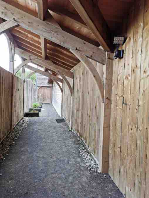 A gravel pathway is framed by wooden beams and fencing, providing a sheltered passageway. The natural wood tones of the structure complement the rustic ambiance, while outdoor lighting fixtures are mounted for visibility.