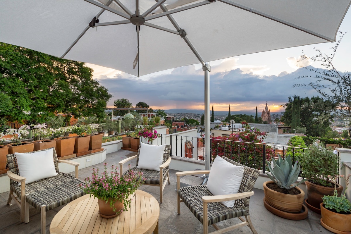 An outdoor seating area is complemented by a large umbrella and comfortable chairs. Potted flowers add vibrancy to the space, with breathtaking views of the surrounding landscape and a sunset in the distance, enhancing the tranquil setting.