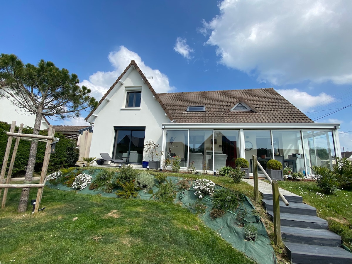A contemporary villa is presented with a large glass front, allowing for abundant natural light. The surrounding garden features various greenery and flowers, while a manicured lawn leads to a set of steps. The sloped roof and large windows contribute to the home's modern appeal.