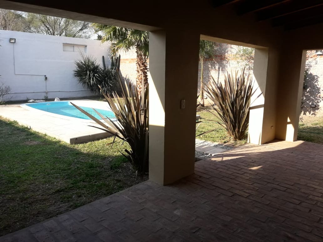 A view of the outdoor space is shown, featuring a swimming pool surrounded by greenery. The area is framed by two supporting columns, with sunlight casting shadows on the brick floor. Low-maintenance plants add a natural touch to the setting.