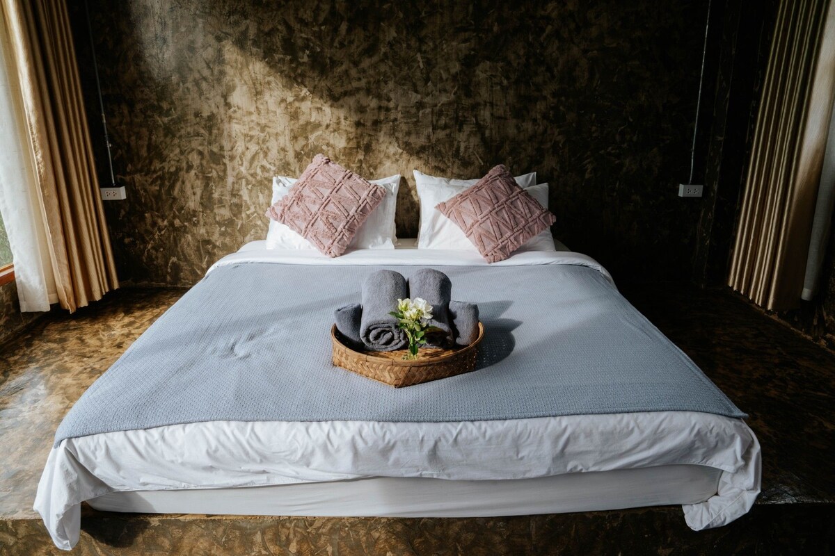 A spacious bed is centered against a textured wall, topped with two decorative pillows. Soft grey linens and a light blanket are complemented by a tray holding rolled towels and a small flower arrangement, enhancing the calming aesthetic of the room.