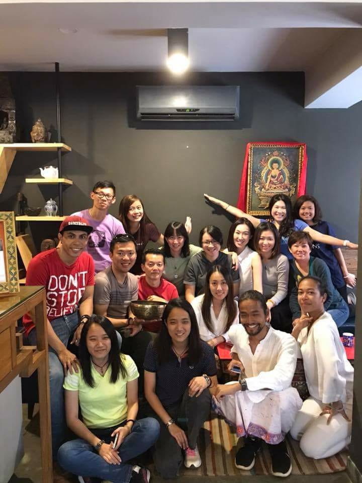 A group of diverse individuals is seated together in a cozy indoor space. Cultural decor is visible on the walls, including a religious artwork. Soft lighting creates a calm environment, while furnishings promote a comfortable gathering for social interaction.