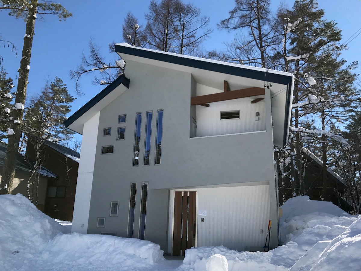 A modern chalet exterior is presented, featuring a clean design with large vertical windows allowing natural light. The building is surrounded by snow, while pine trees are visible in the background. The entrance is marked by double wooden doors and contemporary architectural elements.