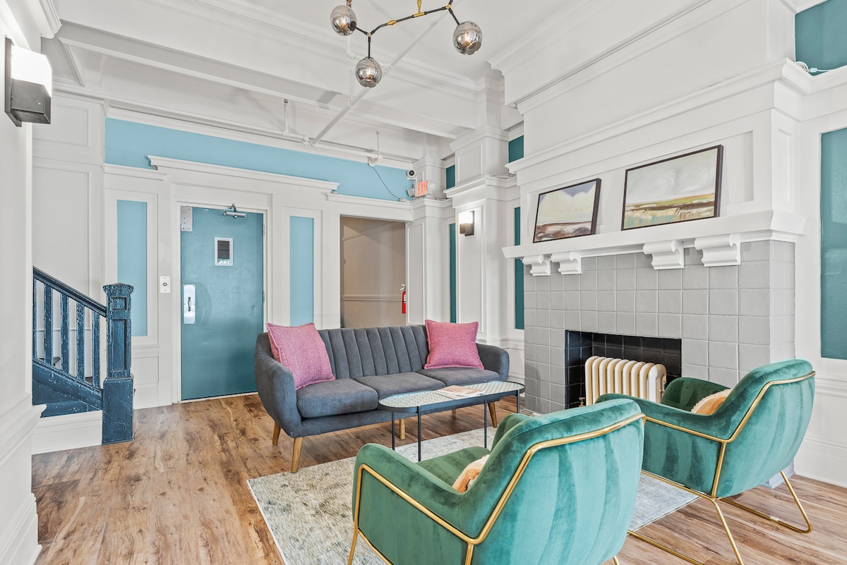 A welcoming lounge area features a modern gray sofa and two stylish teal chairs around a rectangular coffee table. Light-colored hardwood floors complement the bright blue and white walls, while a decorative fireplace adds character. A minimalistic chandelier hangs overhead.