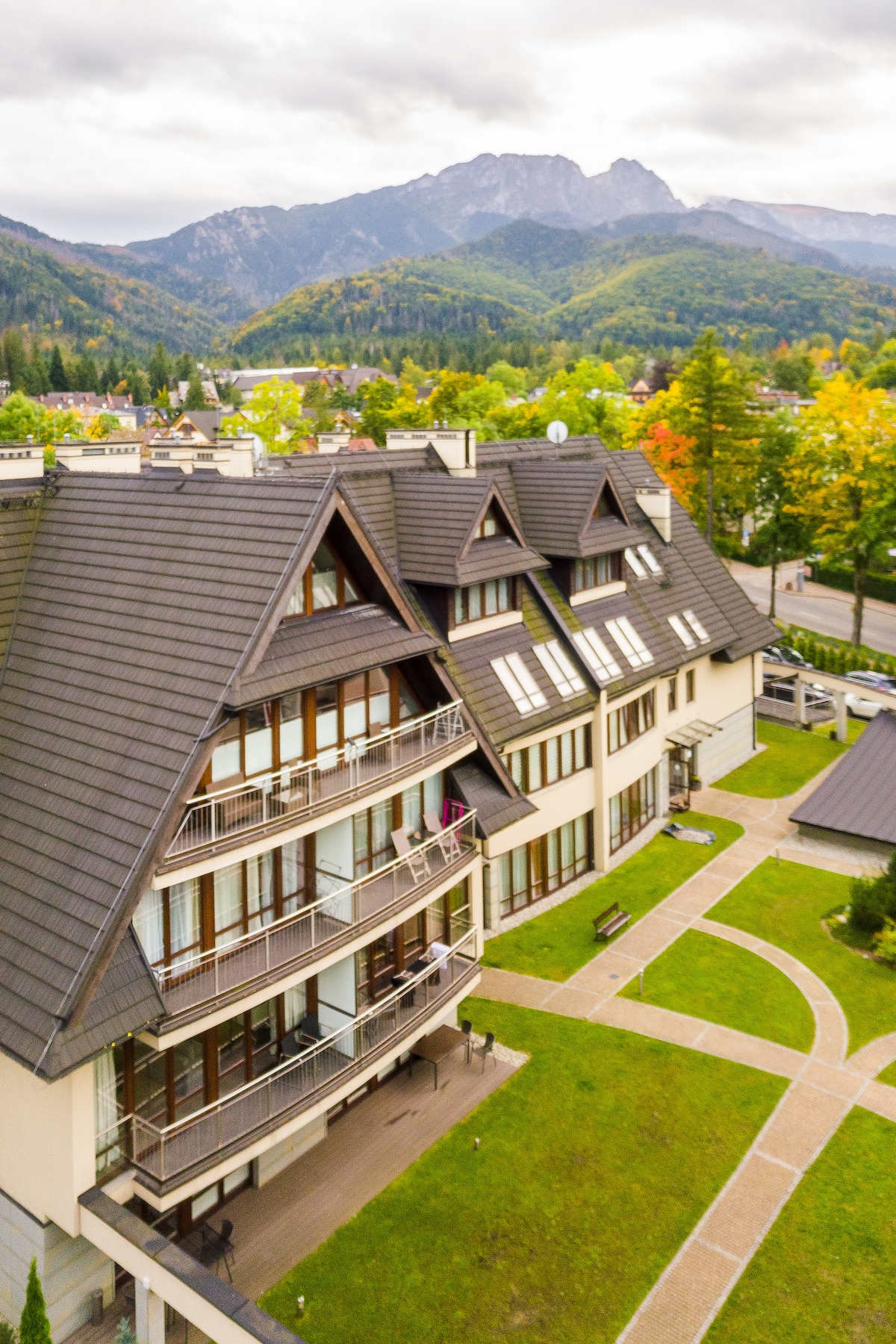 An aerial view captures the modern building nestled in the mountainous landscape. The structure features multiple angles and balcony areas, with neatly landscaped green lawns and pathways. Trees displaying autumn colors frame the scene, enhancing the serene mountain backdrop.