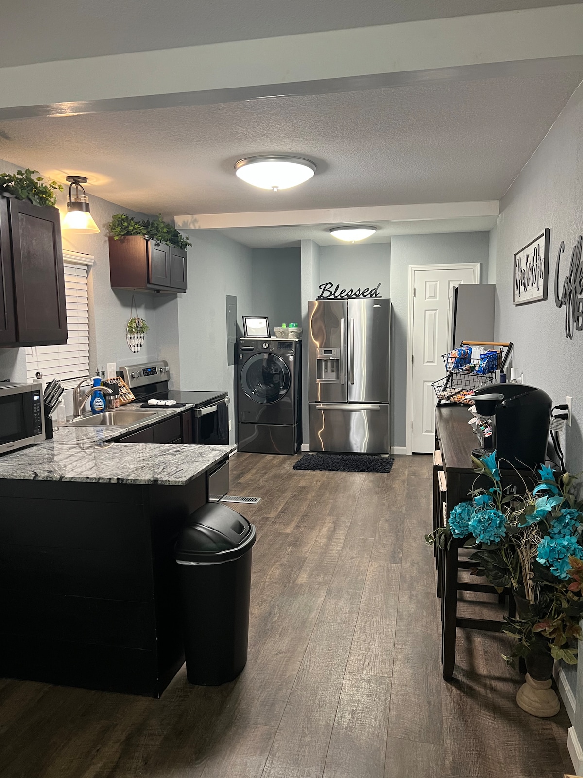 The kitchen features dark cabinetry and a granite countertop with a sink. Stainless steel appliances, including a refrigerator and washer/dryer, are present. A coffee station and decorative items enhance the workspace, while a door leads to another room. Soft lighting adds to the ambiance.