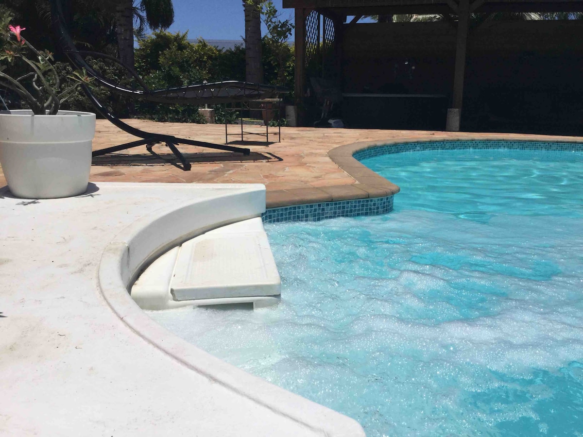 A clear swimming pool is highlighted with a gentle slope leading into the water. The pool's surface reflects sunlight, while a surrounding patio features terra cotta tiles. In the background, a covered area with seating and lush greenery is visible, creating a serene outdoor space.