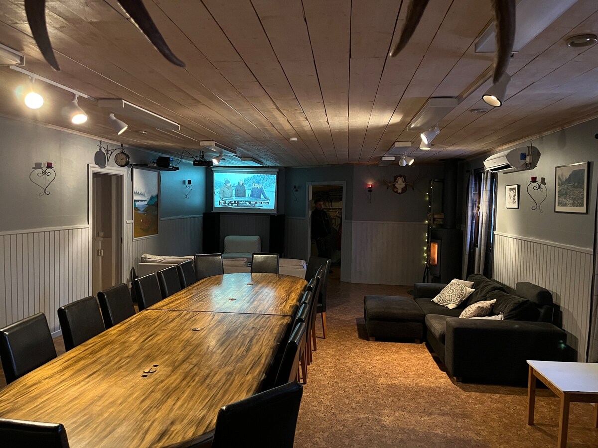 A spacious communal area is shown, featuring a large wooden table surrounded by black chairs. A projection screen is mounted on the wall, and cozy seating is arranged nearby. Soft lighting highlights the wooden ceiling and enhances the inviting atmosphere of the room.