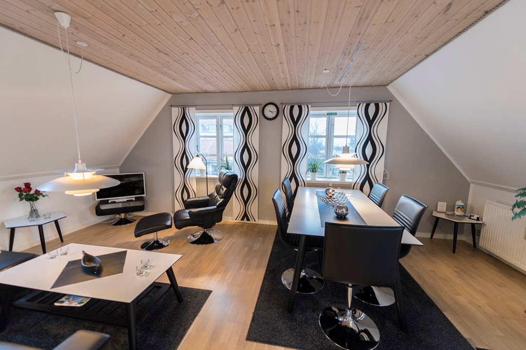 The living room features stylish furnishings, including a black dining table surrounded by sleek chairs. A comfortable lounge chair is positioned nearby, while large windows provide natural light. Decorative curtains complement the modern design, and a wall-mounted television is visible in the background.