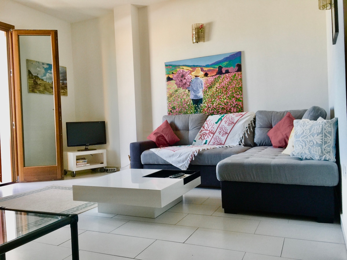 A spacious living area features a large sectional sofa adorned with decorative pillows. A modern coffee table sits at the center, and light streams in through sliding glass doors that lead to the terrace. A vibrant piece of artwork decorates the wall.