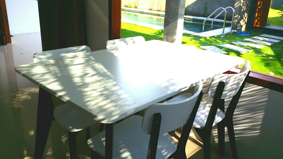 A dining table with six chairs occupies the center of the room, positioned near large windows. Natural light filters through, casting intricate shadows on the white surface. In the background, a swimming pool and lush green lawn are visible, enhancing the space's serene ambiance.
