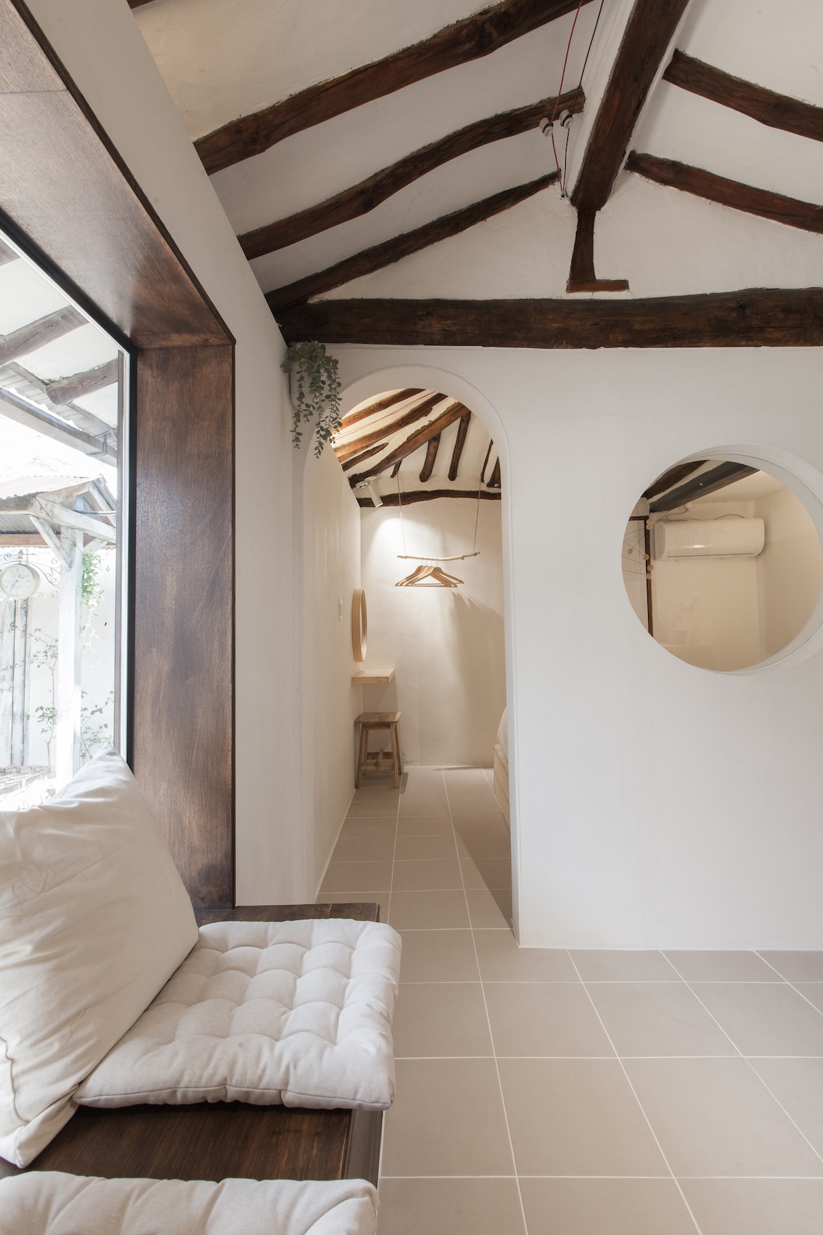A cozy interior space features a bench with soft cushions beneath a large window. Natural light illuminates the narrow corridor leading to the bedroom, showcasing the wooden ceiling beams and an oval mirror on the wall.