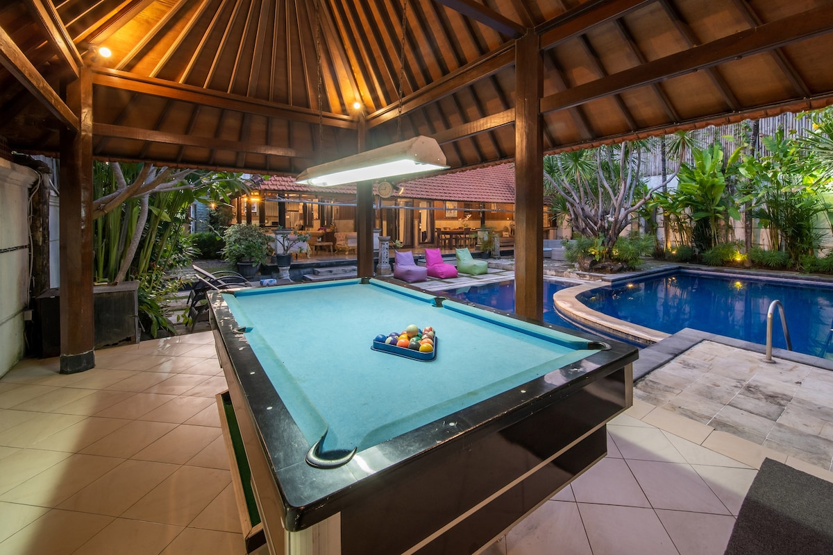 An outdoor entertainment area features a billiards table positioned beneath a traditional roof. Lush greenery surrounds the space, with a shimmering pool visible in the background, enhancing the inviting ambiance. Colorful seating can be seen nearby, creating a relaxing social environment.