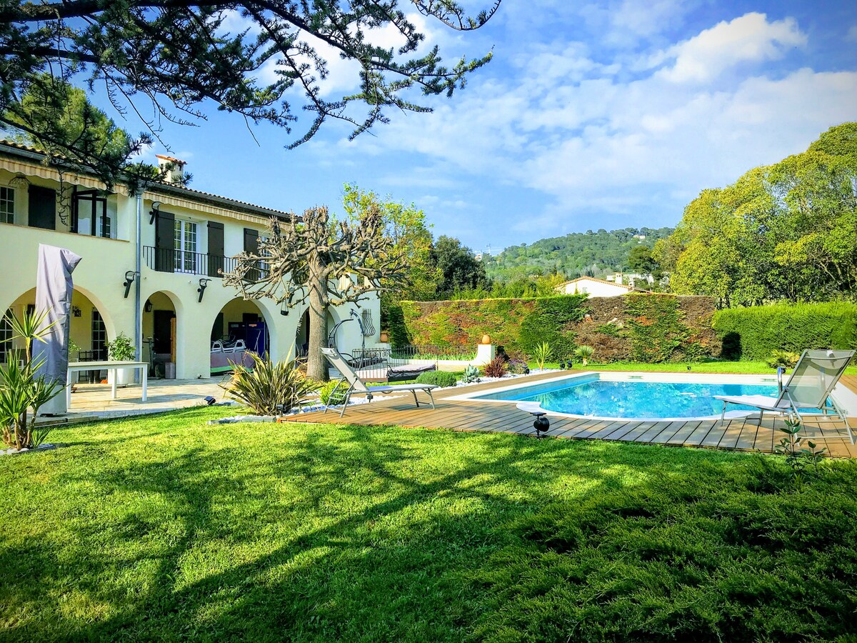 A serene outdoor space features a light blue swimming pool surrounded by wooden decking. Lush green grass and a variety of trees enhance the setting. The two-story villa stands in the background, characterized by arched windows and a balcony overlooking the garden.