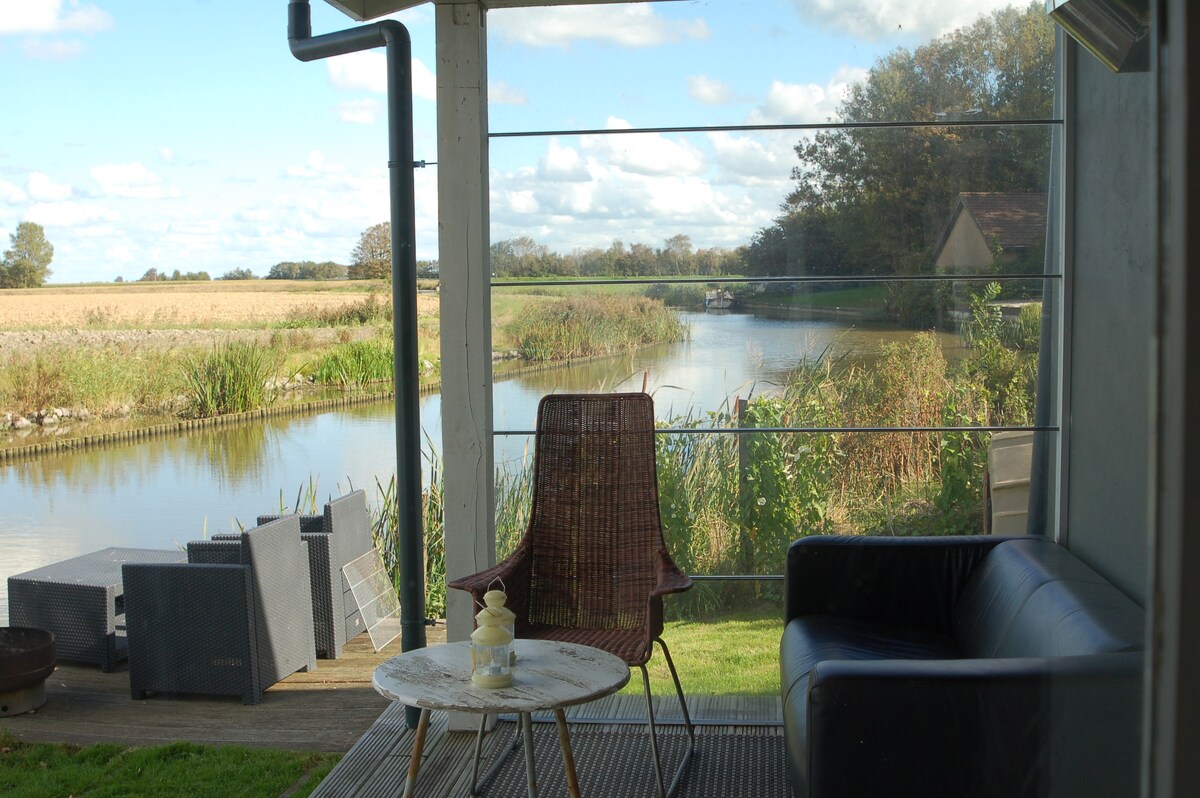 A serene outdoor seating area is showcased, featuring a round table and a comfortable chair, with a second chair visible nearby. The view overlooks a calm waterway, surrounded by grassy banks and distant fields, under a blue sky with scattered clouds.