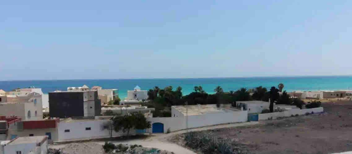 A scenic view captures a vibrant coastline with a clear blue ocean meeting the sky. Residential buildings with varying architectural styles are visible, interspersed with palm trees and coastal vegetation, creating a serene atmosphere. The landscape features sandy areas adjacent to the structures.