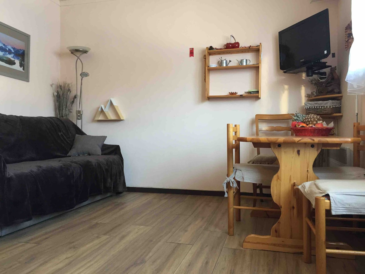A cozy living area features a dark sofa alongside a light wooden dining table with multiple chairs. A wall-mounted television and shelving display decorative items. Natural light enters through a nearby window, enhancing the warm space.
