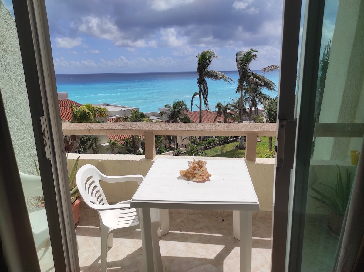 A private balcony offers an unobstructed view of the ocean, where turquoise waters meet a clear sky. A light-colored table is placed at center, complemented by a white plastic chair. Palm trees sway gently in the breeze, enhancing the coastal ambiance.