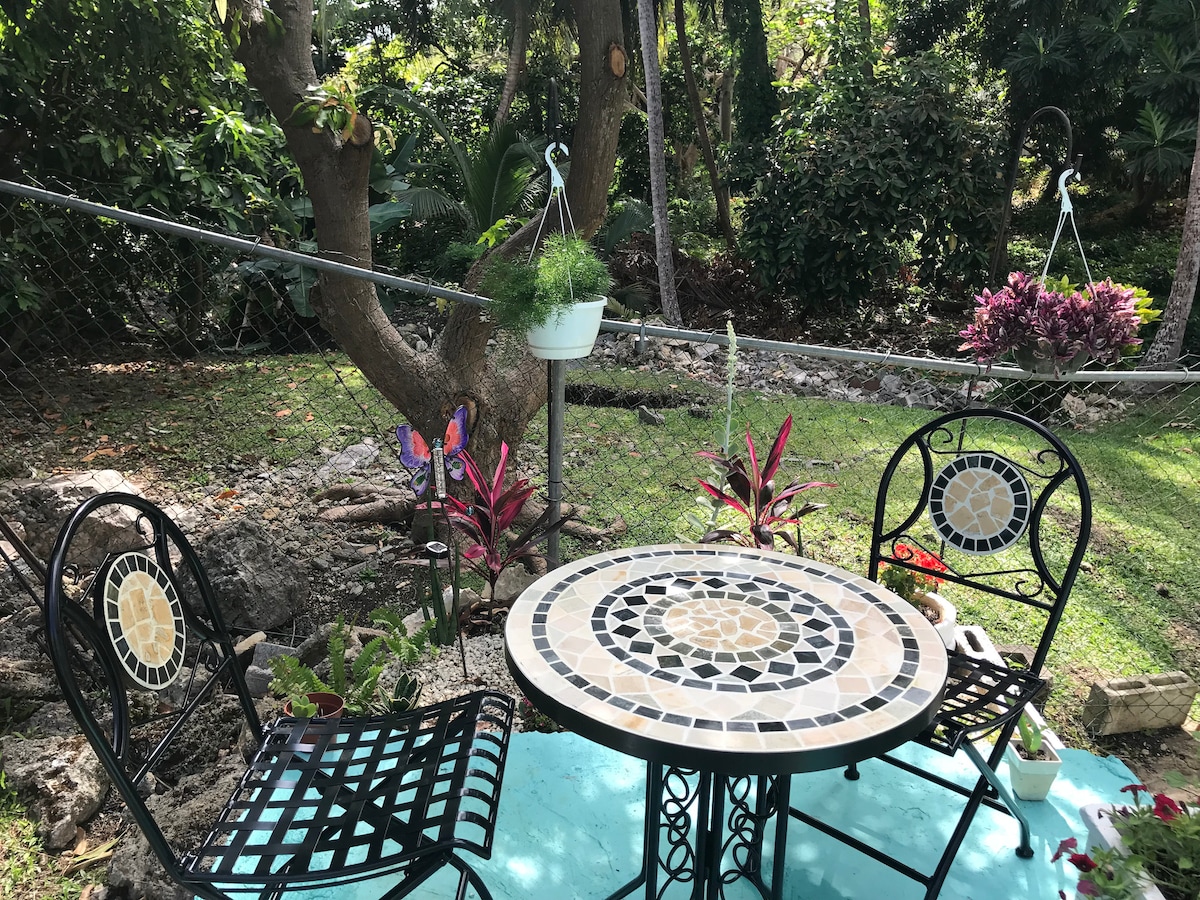 A small outdoor sitting area features a round mosaic table surrounded by two black metal chairs. Greenery serves as the backdrop, with various plants and a decorative hanging planter adding vibrant colors to the tranquil garden space.