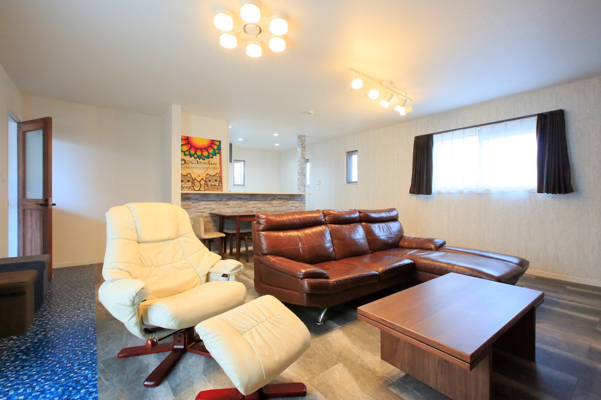 A spacious living area features a large brown leather sectional sofa and a matching lounge chair. A wooden coffee table is placed in front of the seating. Soft lighting is provided by modern fixtures above, while natural light enters through a window with light curtains.