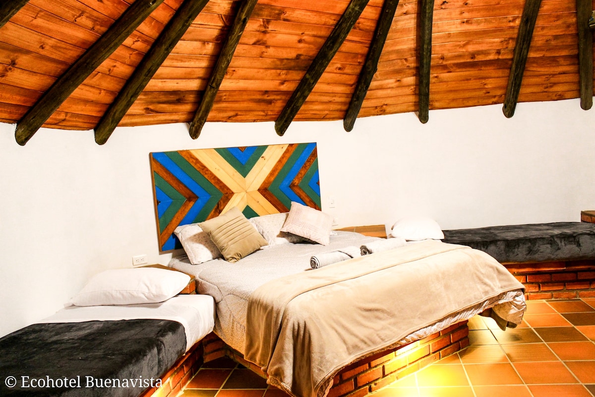 A spacious sleeping area is highlighted by a large bed with a textured blanket. Two additional beds are positioned around the room. The ceiling features exposed wooden beams, and a colorful headboard adds a distinctive touch to the neutral walls.