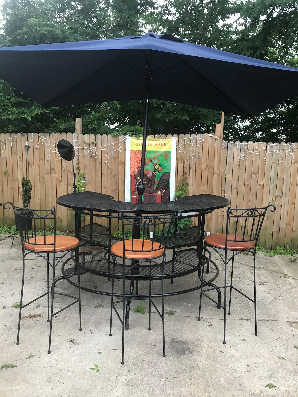 An outdoor seating area features a round wrought iron table shaded by a large umbrella. Four metal chairs, each with cushioned seats, surround the table. A colorful art piece is visible in the background, alongside a wooden fence adorned with decorative lights.