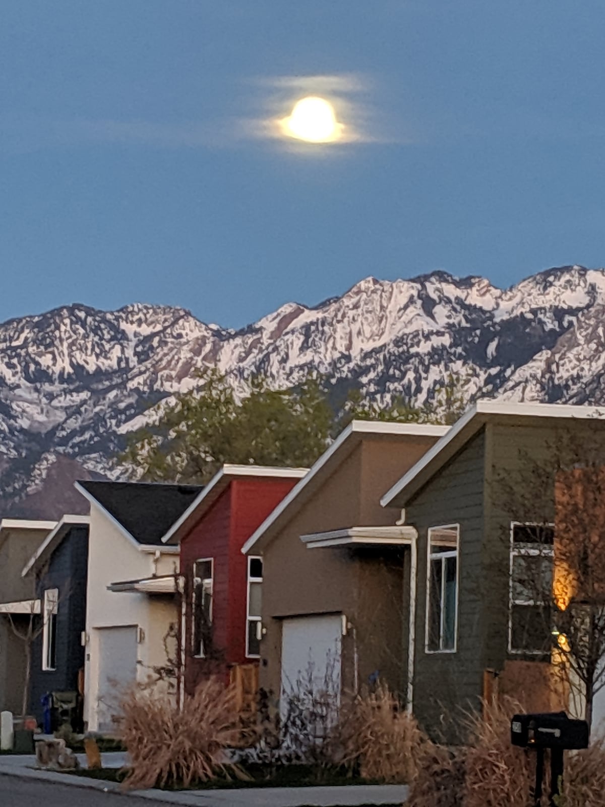 A serene evening scene showcases a cluster of modern homes with varied facades, framed by dramatic snow-capped mountains under a clear sky. The moon glows softly overhead, illuminating the neighborhood's quiet ambiance.